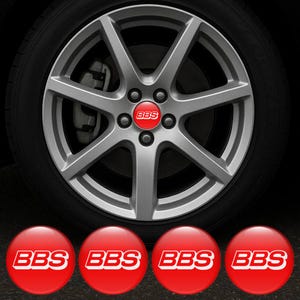 以下が含まれることがあります： 白い「BBS」の文字が入った赤いセンターキャップが付いたシルバーの合金ホイール。ホイールは黒いタイヤに取り付けられています。4つの追加の赤いセンターキャップには「BBS」が表示されています。