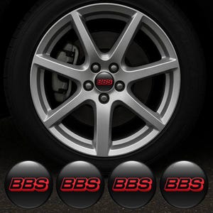 以下が含まれることがあります： 黒いタイヤが付いたシルバーの合金ホイール。ホイールはマルチスポークデザインで、赤い文字「BBS」のセンターキャップが付いています。赤い「BBS」ロゴが付いた4つの追加の黒いセンターキャップがホイールの下にあります。