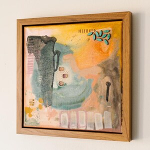 May include: A square abstract painting in a light wood frame featuring a textured composition of yellow, orange, and teal paint. The artwork includes a dark charcoal vertical shape, small turquoise leaf-like accents, and translucent beads.
