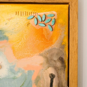 May include: A framed canvas painting featuring a yellow, orange, and light blue abstract background. The artwork includes a cluster of small, oval-shaped turquoise beads attached to the surface, alongside a series of vertical brown stitched lines.