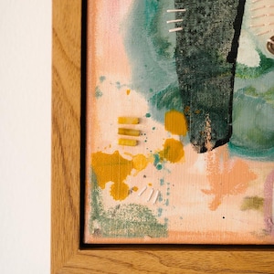 May include: A framed mixed media artwork on canvas featuring abstract layers of peach, teal, and dark green paint. The composition includes yellow paint splatters, small horizontal yellow rectangular accents, and white stitched thread details.