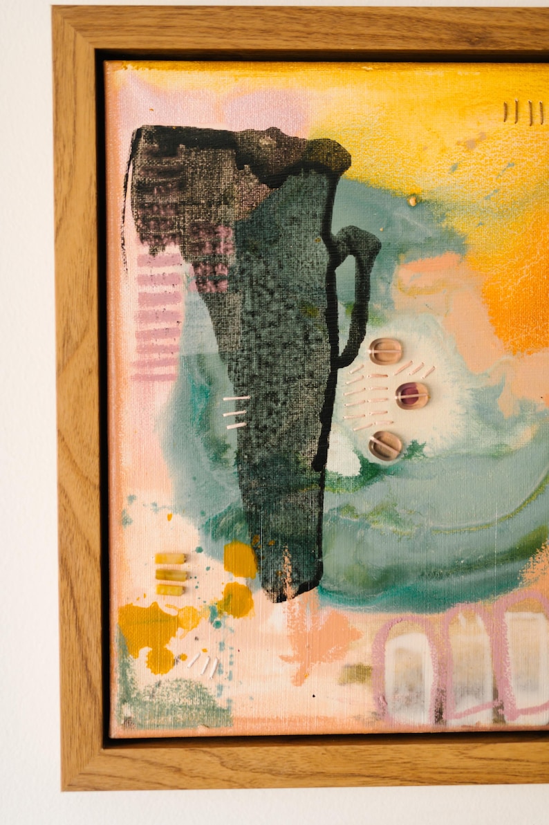 May include: A framed mixed media artwork featuring a central dark green textured shape resembling a vessel against a background of yellow, peach, and teal paint washes. The piece includes small circular translucent beads and stitched thread details.