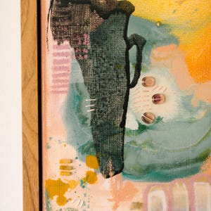 May include: An abstract painting in a light wood frame featuring a dark green textured central shape, yellow paint splatters, and soft pink and blue washes. The composition includes small stitched details and three translucent circular embellishments.