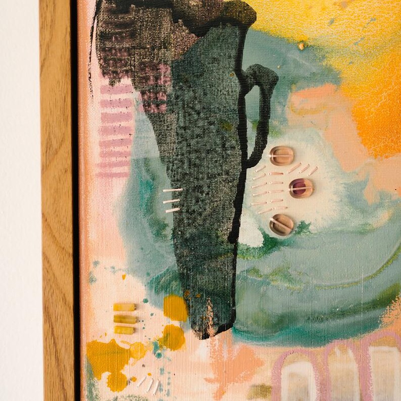 May include: An abstract painting in a light wood frame featuring a dark green textured central shape, yellow paint splatters, and soft pink and blue washes. The composition includes small stitched details and three translucent circular embellishments.