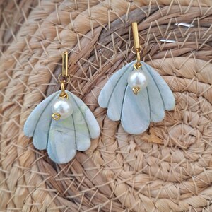 Peut inclure: Une paire de boucles d'oreilles pendantes composées de pendentifs en argile en forme d'éventail aux motifs marbrés bleu clair et blanc. Chaque pièce est ornée d'une perle blanche et suspendue à une barre métallique dorée.