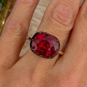 Puede incluir: Un anillo con una gran piedra preciosa de color rojo intenso, forma ovalada y tallada en facetas, engastada en un bisel de metal oscuro. La piedra muestra un tono carmesí vibrante con múltiples facetas reflectantes, sobre una banda dorada fina.