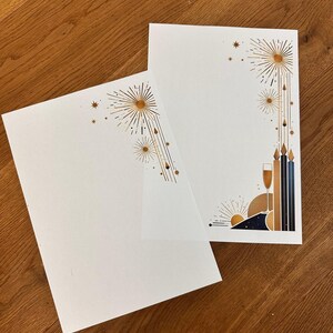 May include: Two white stationery cards with gold and navy blue art deco designs. One card features a sunburst design, while the other has a champagne glass and candle motif. The cards are on a wooden surface.