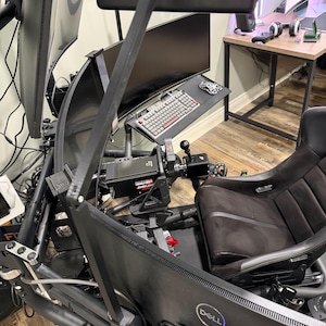 Op de afbeelding: Een zwarte racesimulatoropstelling met meerdere gebogen monitoren, een stuurwiel en een racestoel. Een toetsenbord en muis staan op een zijtafel. Het Dell-logo is zichtbaar op een van de monitoren.