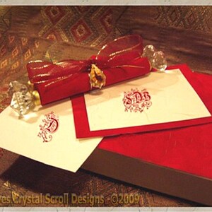 May include: A red and gold scroll with a crystal embellishment, resting on a red and white stationery set with a monogram. The stationery set includes two sheets of paper with red monograms and a red envelope.