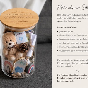 Op de afbeelding: Een doorzichtige glazen pot met een rond houten deksel gegraveerd met de tekst P&Auml;DAGOGISCHE Notfallration F&Uuml;R DIE BESTE DER GANZEN WELT. De pot is gevuld met een kleine pluche teddybeer, een dennenappel, opgerolde papieren briefjes en kaartjes met het woord Danke.