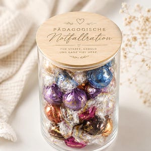 Op de afbeelding: Een doorzichtige glazen pot gevuld met diverse Lindt Lindor chocoladetruffels in kleurrijke folieverpakkingen. De pot heeft een rond houten deksel met de gravure P&Auml;DAGOGISCHE Notfallration, F&Uuml;R ST&Auml;RKE, GEDULD UND GANZ VIEL HERZ en decoratieve hartjes.
