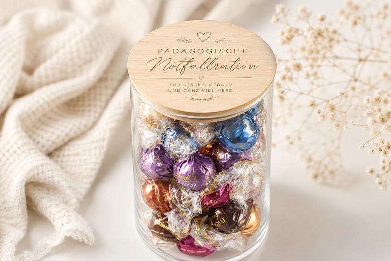 Op de afbeelding: Een doorzichtige glazen pot gevuld met diverse Lindt Lindor chocoladetruffels in kleurrijke folieverpakkingen. De pot heeft een rond houten deksel met de gravure P&Auml;DAGOGISCHE Notfallration, F&Uuml;R ST&Auml;RKE, GEDULD UND GANZ VIEL HERZ en decoratieve hartjes.