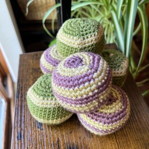 May include: A collection of six crocheted spheres in shades of green, purple, and cream. The handmade spheres are stacked on a wooden surface, showcasing the texture and color variations of the yarn. The spheres are a unique decorative item.