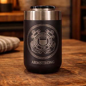 May include: A black and silver insulated tumbler with the United States Coast Guard emblem and the name "ARMSTRONG" engraved on it. The tumbler is cylindrical and sits on a wooden surface.