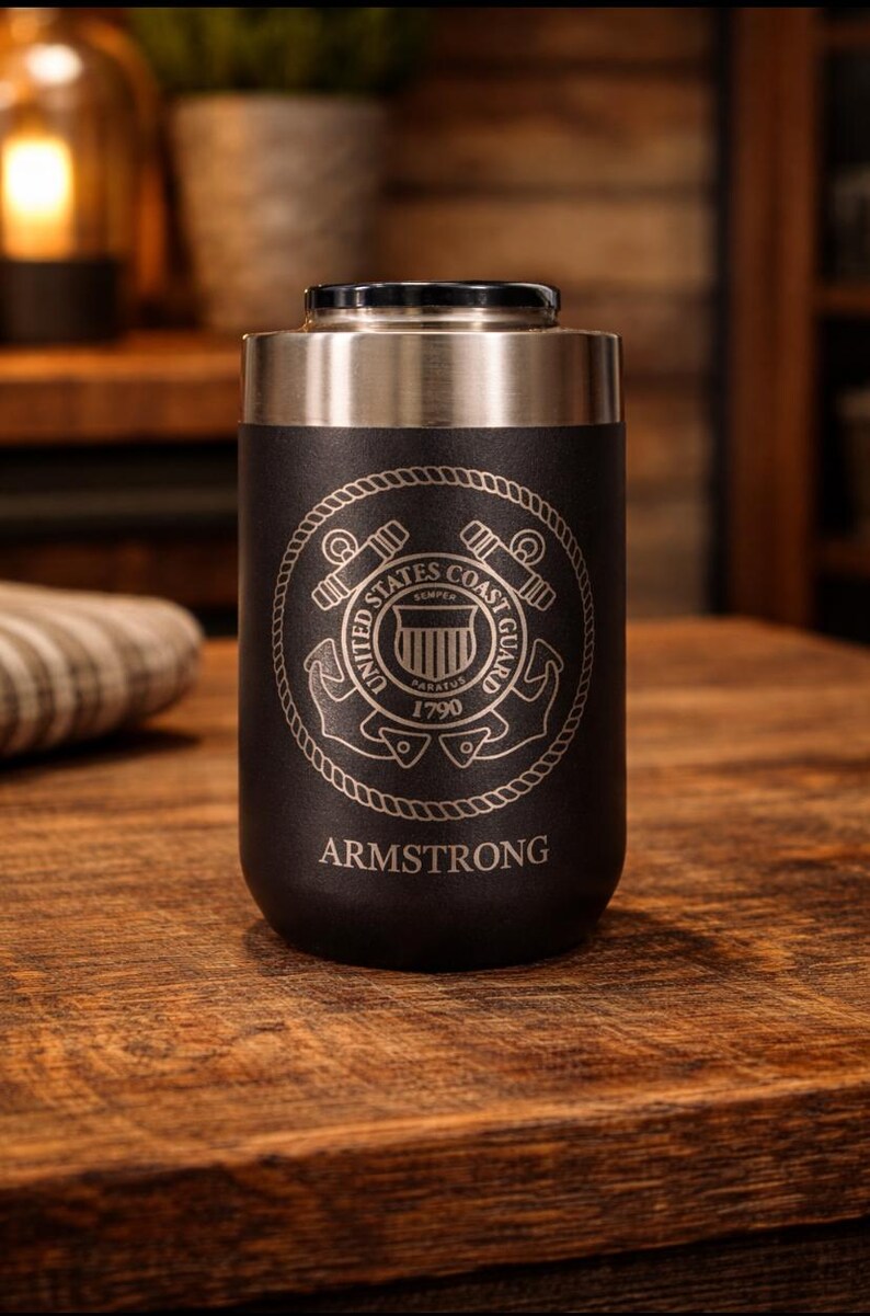 May include: A black and silver insulated tumbler with the United States Coast Guard emblem and the name "ARMSTRONG" engraved on it. The tumbler is cylindrical and sits on a wooden surface.
