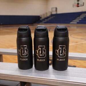 May include: Three black water bottles with a silver logo and the name "PLEICH" are displayed on a bench. The bottles have a flip-top lid and are cylindrical in shape. The background shows a basketball court.