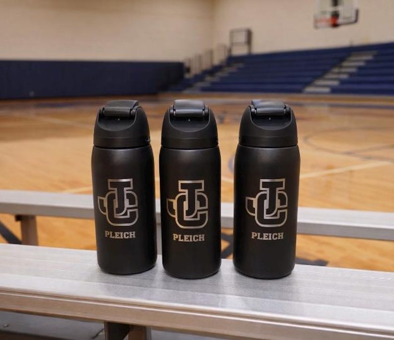 May include: Three black water bottles with a silver logo and the name "PLEICH" are displayed on a bench. The bottles have a flip-top lid and are cylindrical in shape. The background shows a basketball court.