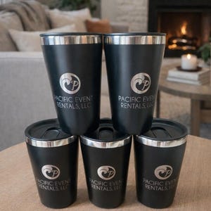 May include: A stack of six black insulated tumblers with silver rims and lids. Each tumbler has the "Pacific Event Rentals, LLC" logo printed in silver. The tumblers are arranged on a round wooden table.
