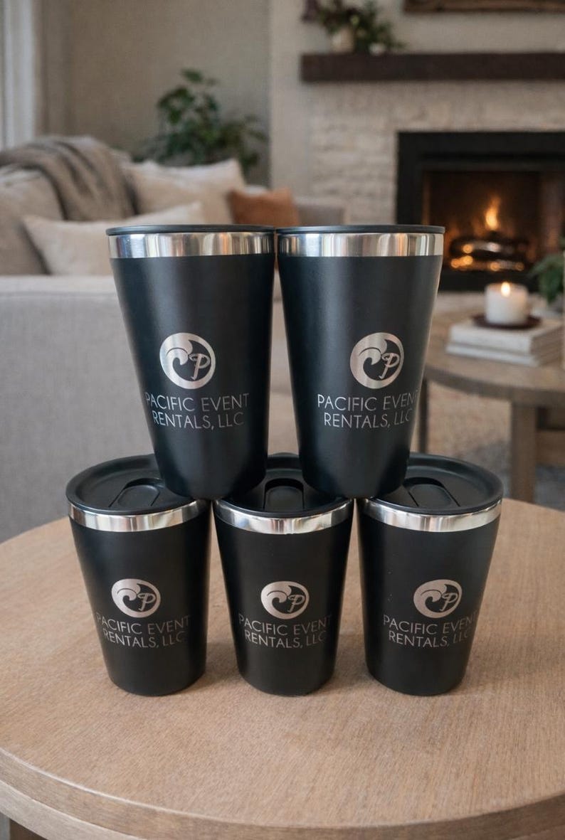 May include: A stack of six black insulated tumblers with silver rims and lids. Each tumbler has the "Pacific Event Rentals, LLC" logo printed in silver. The tumblers are arranged on a round wooden table.