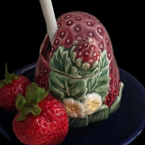 May include: A ceramic strawberry-shaped jar with a lid, featuring a textured red surface with small indentations, green leaves, and white flowers. A white ceramic spoon rests inside the jar. Two fresh red strawberries sit beside the jar.