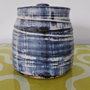 May include: A ceramic jar with a lid, displaying a blue and white striped design. The jar has a rounded form and a small, circular lid. The design incorporates dark blue and white horizontal stripes, giving a textured look. The jar is likely handmade, with a height of approximately 15 cm.