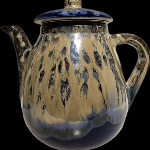 May include: A ceramic teapot with a lid, featuring a speckled glaze in shades of tan, blue, and brown. The teapot has a curved spout and a handle. The lid is a darker blue. The teapot is designed for brewing tea.