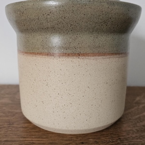 May include: A ceramic jar with a lid, featuring a speckled, two-tone design. The upper portion and lid are a speckled grey, while the lower section is a speckled cream colour. A thin, reddish-brown band separates the two colours.