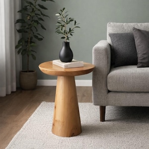 Puede incluir: Una mesa auxiliar de madera clara con una parte superior redonda y una base cónica. Un jarrón negro con vegetación y un libro están sobre la mesa. La mesa está junto a un sofá gris y una planta en maceta. La mesa mide unos 60 cm de alto.