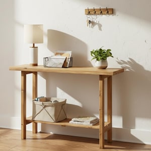 Può includere: Una consolle in legno marrone chiaro con un ripiano inferiore. Una lampada beige, un cestino di filo metallico, una piccola pianta in vaso e un portachiavi sono sul tavolo. Un cestino di stoffa beige e libri sono sul ripiano inferiore.