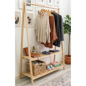Op de afbeelding: Een lichtgekleurde houten kledingrek met twee planken. De bovenste plank bevat kleding op houten hangers. De onderste planken tonen een gevlochten mand, boeken en schoenen. Een potplant staat rechts.