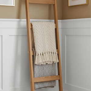 May include: A wooden blanket ladder with two decorative blankets. The top blanket is cream-coloured with a chevron pattern and fringe. The bottom blanket is grey with a floral pattern. The ladder is made of light-coloured wood.