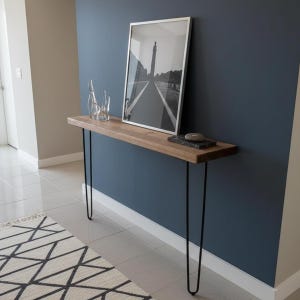 Puede incluir: Una consola de madera con patas de horquilla negras. Una fotografía en blanco y negro enmarcada y botellas de vidrio decorativas están sobre la mesa. La mesa está contra una pared azul oscuro en un pasillo.