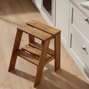 May include: A two-step wooden step stool with a natural finish. The stool has a rectangular top and a lower step, supported by angled legs. The wood grain is visible, and the stool is designed for reaching higher shelves or cabinets.