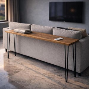 Puede incluir: Una mesa consola larga y estrecha de madera con acabado natural, sostenida por patas negras de horquilla. La mesa está colocada detrás de un sofá gris y contiene una pila de libros y un teléfono inteligente. La mesa mide unos 152 cm de largo.