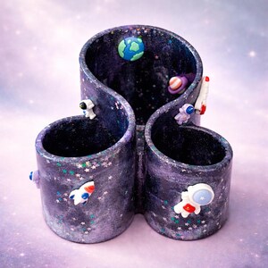 Könnte beinhalten: Ein skurriler Schreibtisch-Organizer im Weltraum-Thema. Das lila-schwarze Harzstück hat drei zylindrische Fächer, die durch ein wellenförmiges Design verbunden sind. Verziert mit Miniatur-Astronauten, Raketen, Planeten und sternförmigem Glitzer.