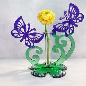 May include: A decorative vase with a yellow flower. The vase has a clear glass tube for the flower, supported by a green base with purple butterfly accents. The butterflies and base have a decorative design.