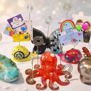 May include: A selection of decorative photo holders in the shapes of a crocodile, snail, octopus, and reindeer. Each has a spiral wire for holding photos or notes. The holders are brightly coloured and whimsical.