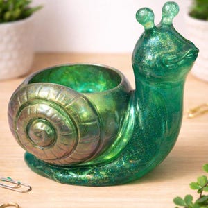 Puede incluir: Una maceta decorativa en forma de caracol verde con un acabado brillante. La concha del caracol tiene un diseño en espiral y el cuerpo tiene una textura suave y brillante. La maceta está diseñada para contener pequeñas plantas o elementos decorativos.