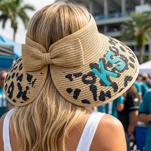 May include: A straw visor with a leopard print design and a large bow. The visor has the letters "JAX" in teal. The person is wearing a white dress and is at an outdoor event.