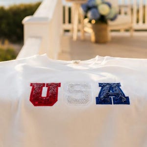 May include: A white long-sleeve shirt with the letters "USA" in red, white, and blue. The shirt is displayed on a white railing with a beach and ocean in the background. The shirt is made of a soft material.