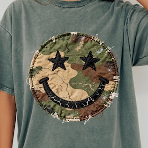 May include: A sage green t-shirt featuring a large, circular patch with a smiley face design. The smiley face has star eyes and a stitched, camouflage pattern. The shirt has short sleeves and a relaxed fit.