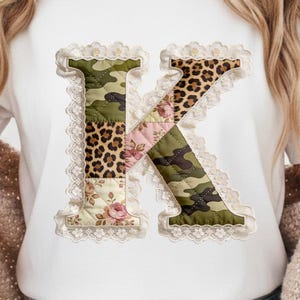 Può includere: T-shirt bianco con una grande lettera "K" decorativa. La lettera è composta da vari tessuti fantasia, tra cui camouflage, stampa leopardata e disegni floreali, il tutto bordato da pizzo bianco.