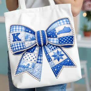 May include: White tote bag with a large, decorative blue bow. The bow features a patchwork design with various blue patterns, including gingham and star motifs, and is outlined with white pearls. The letter "K" is visible on the bow.