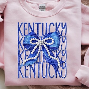 May include: A light pink sweatshirt with the word "Kentucky" in blue script repeated vertically. A large blue bow with white pearl accents is centered on the front. The sweatshirt has a crew neck and long sleeves.