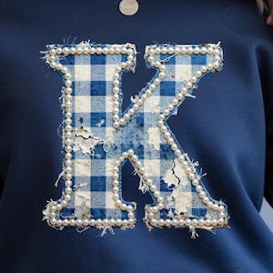 Könnte beinhalten: Marineblaues Sweatshirt mit einem großen Buchstaben "K"-Design. Der Buchstabe ist mit einem blau-weiß karierten Muster gefüllt und mit Kunstperlen umrandet. Das Design hat eine Used-Optik im Vintage-Stil.