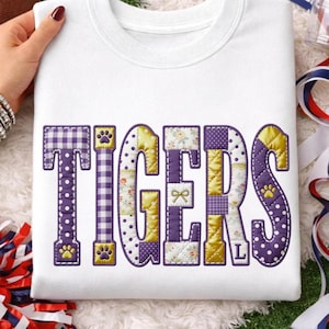 Peut inclure: Sweat-shirt blanc avec le mot "TIGERS" dans un motif patchwork. Les lettres sont violettes, dorées et blanches, avec des empreintes de pattes et des motifs floraux. La lettre "L" est visible. Le sweat-shirt est tenu par une main.
