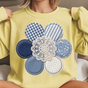May include: A yellow sweatshirt featuring a large flower design. The flower is composed of blue and white patterned fabric petals, including gingham, stripes, and floral prints, surrounding a white lace doily center. The petals are stitched onto the shirt.