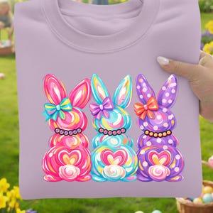 May include: A light purple sweatshirt featuring a colorful graphic of three Easter bunnies. Each bunny has a bow, a heart-shaped belly, and a candy-like design in pink, blue, and purple. The bunnies are on a light purple background.