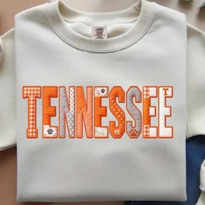 May include: Cream-colored sweatshirt with the word "TENNESSEE" in orange, gray, and white patchwork lettering. The letters feature various patterns, including gingham, polka dots, and paw prints. A small tag is visible at the top.