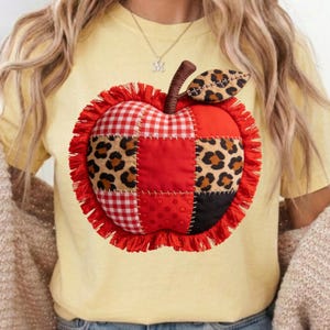 May include: A yellow t-shirt with an appliqued apple design. The apple is made from fabric patches, including red gingham, leopard print, and solid red and black pieces. It has a brown stem, a leopard print leaf, and a red fringe border.